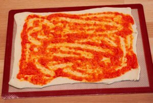 Paprikasauce-fuer-Spanische-Pizza-auf-dem-frischen-Teig-aufgetragen-teig-auf-Silikonmatte ausgerollt