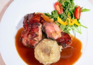 Gegrillte-Schweinshaxe-sous-Vide-mit-Kartoffelknoedel-kleinem-Salat-und-Biersauce-auf-weissem-Teller
