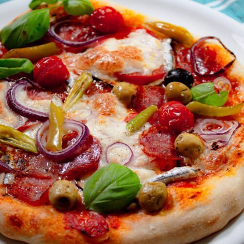 Fertige-Salamipizza-mit-Zwiebeln-Mozerella-Paeperoni-und-Cherrytomaten-auf-weissem-Teller-