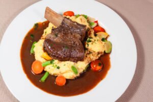 Angus-geschmorte-Short-Rip-auf-Polenta-mit-Gemuese-und-dunklem-Saucenspiegel-auf-weissem-Teller