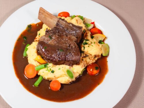 Angus-geschmorte-Short-Rip-auf-Polenta-mit-Gemuese-und-dunklem-Saucenspiegel-auf-weissem-Teller