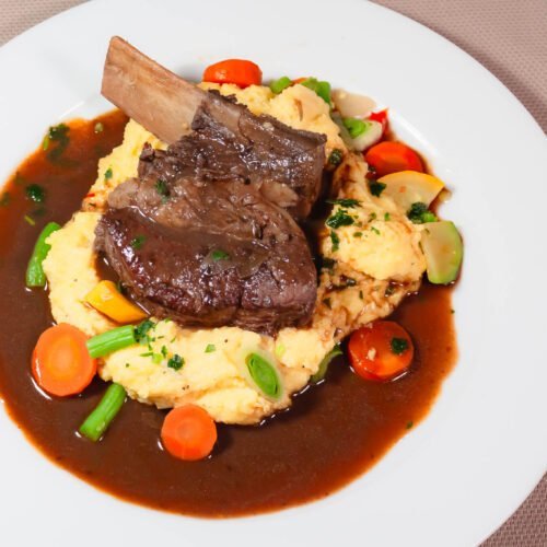 Angus-geschmorte-Short-Rip-auf-Polenta-mit-Gemuese-und-dunklem-Saucenspiegel-auf-weissem-Teller